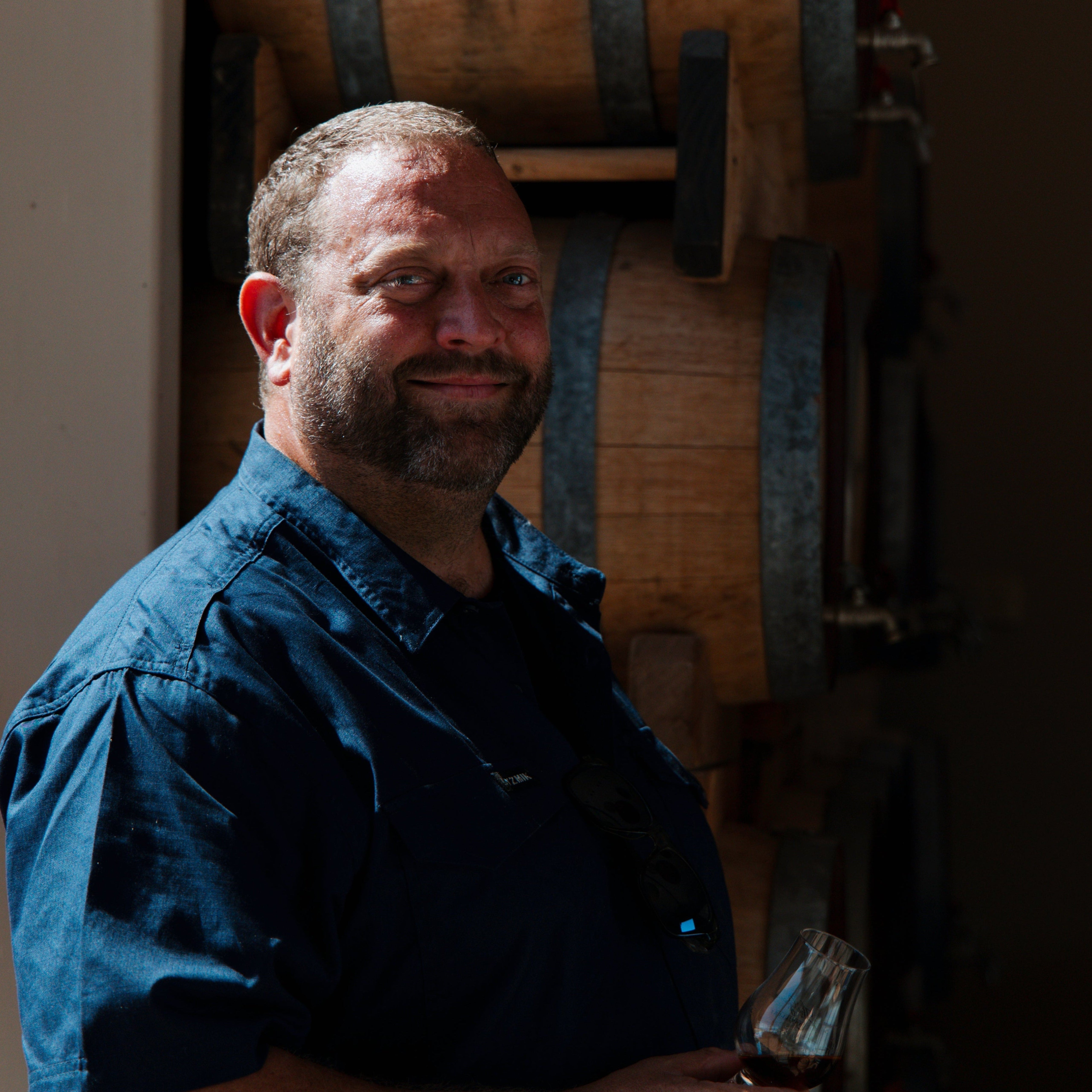 Man in a blue shirt standing in a dimly lit room with wooden barrels in the background